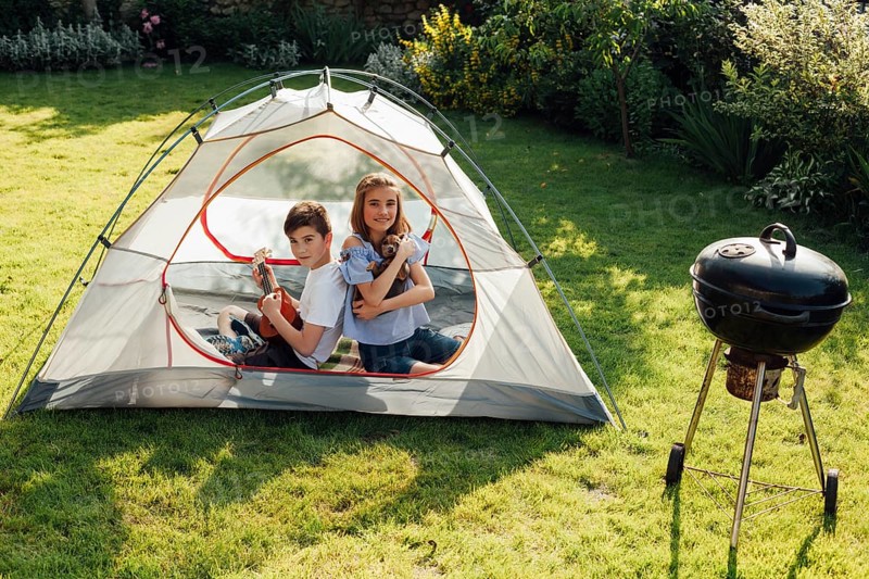 Boy playing ukulele sitting back back his sister tent near barbecue grill