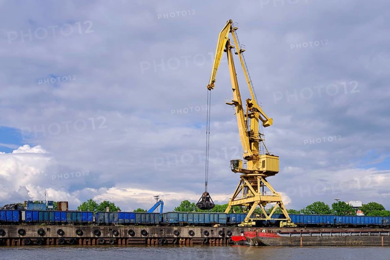 River port crane with clamshell or griper loading coal from open-top gondola car or wagon to river drag boats or barges moored by pier on cloudy day. Problems