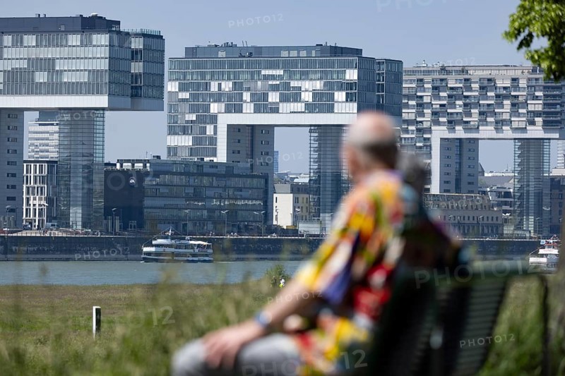 Senior couple sitting on a bench on the banks of the Rhine in front of the crane houses in Cologne's Rheinauhafen harbour on the other bank