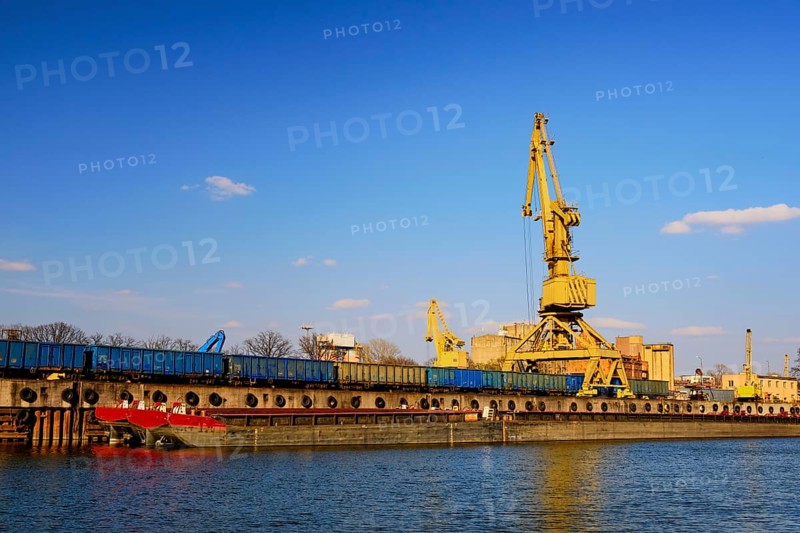 Colorful wide shot of river port crane loading open-top gondola cars on sunny day. Empty river drag boats or barges moored by pier. Empty cars ready for loading. Shipping raw materials