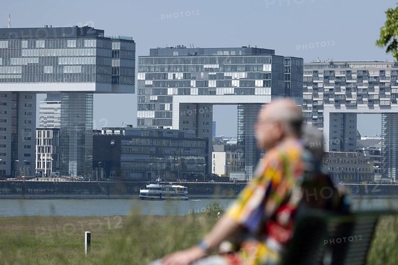 Senior couple sitting on a bench on the banks of the Rhine in front of the crane houses in Cologne's Rheinauhafen harbour on the other bank