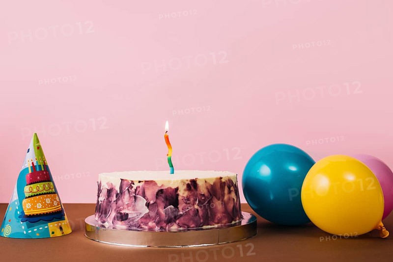Colorful illuminated candle birthday cake with party hat balloons desk against pink background