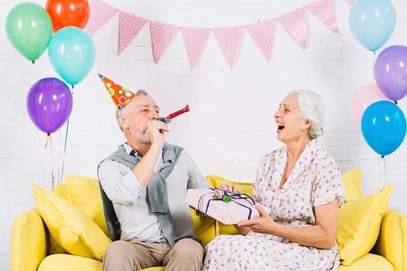 Happy woman with birthday gift looking her husband blowing party horn