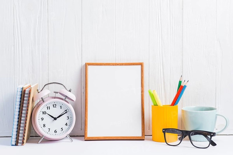 Notebooks alarm clock blank frame pencils holder cup eyeglasses against white wooden wallpaper