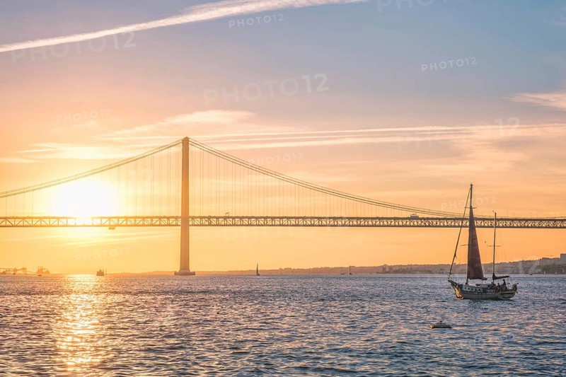 View of 25 de Abril Bridge famous tourist landmark of Lisbon connecting Lisboa and Almada over Tagus river with tourist yacht silhouette at sunset. Lisbon