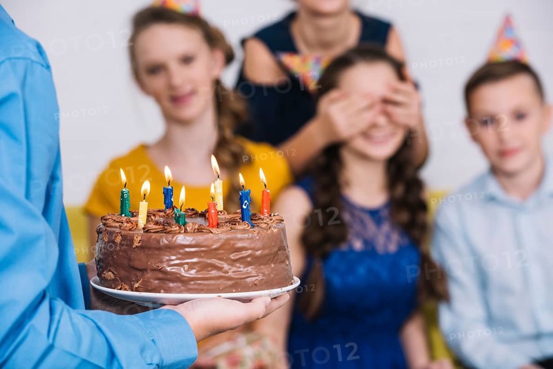 Close up boy s hand bringing chocolate cake birthday girl with covered eyes by her friend