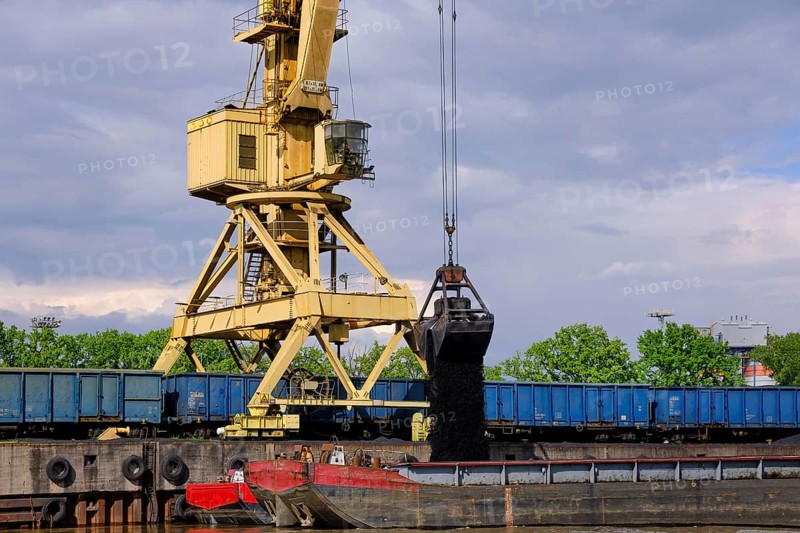 River port crane with clamshell or griper loading coal from open-top gondola car or wagon to river drag boats or barges moored by pier on cloudy day. Problems