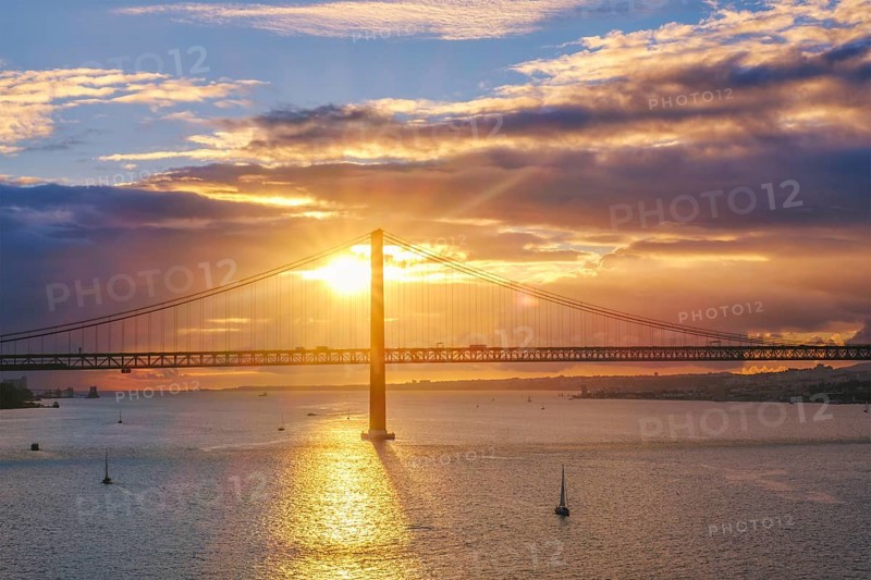 View of 25 de Abril Bridge famous tourist landmark of Lisbon connecting Lisboa and Almada on Setubal Peninsula over Tagus river with tourist yacht boats on sunset. Lisbon