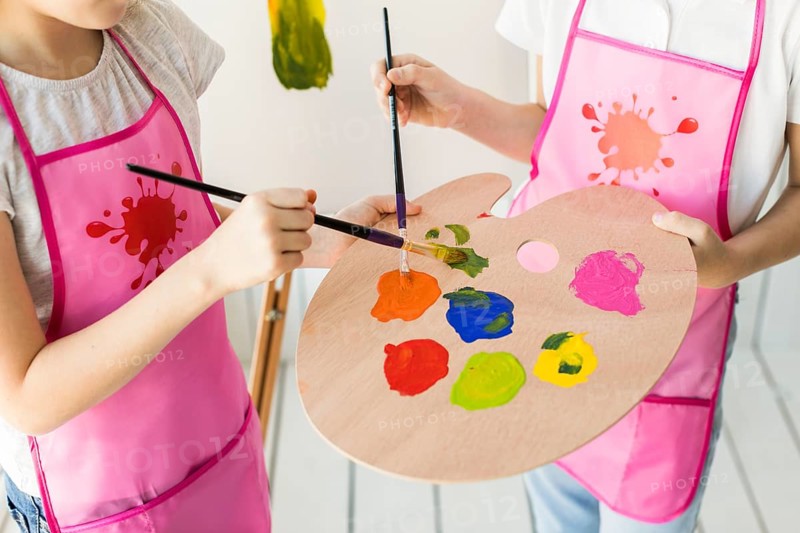 Elevated view two girls same pink apron mixing paint wooden palette with paint brush