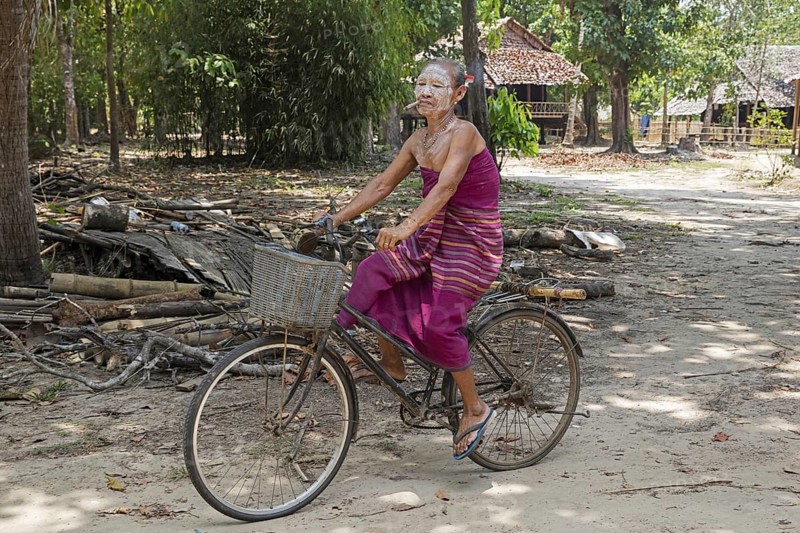 Old Burmese woman wearing thanaka and smoking a cigar while cycling in Kayin village near Hpa-an