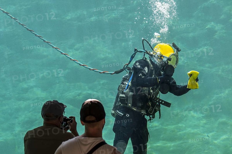 Visitors watching surface-supplied diver equipped with umbilical cleaning glass on inside of huge water tank