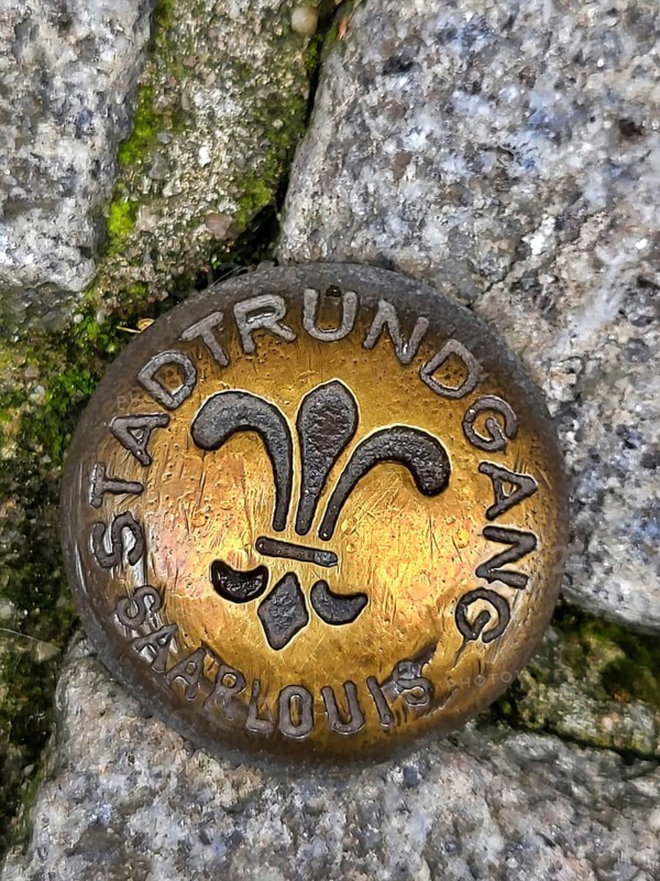 Brass-coloured metal medal embedded in a cobblestone street. The medal is circular and has a fleur-de-lis symbol in the centre. The medal is inscribed with the words Stadtrundgang and Saarlouis around the edge. Saarlouis