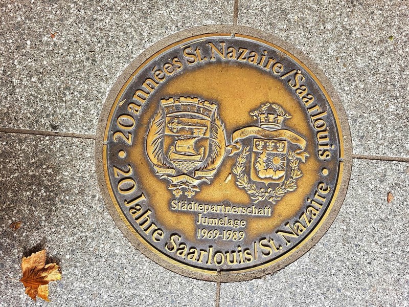 Bronze plaque embedded in a pavement. The plaque is in German and reads 20 years of St Nazaire Saarlouis town twinning jumelage 1969-1989. The plaque has two coats of arms. Saarlouis