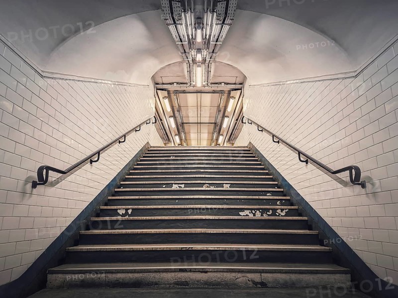 Subway corridor with white tile on walls and glowing lights above a the stairs passage. Symmetrical underground staircase