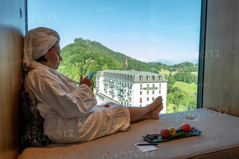 Woman in Bathrobe and Using Her SmartPhone and Enjoy the Window View over Mountain and Building in a Sunny Summer Day in Burgenstock
