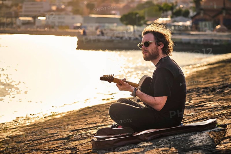 Hipster street musician in black playing electric guitar in the street sitting on pier embankment on sunset