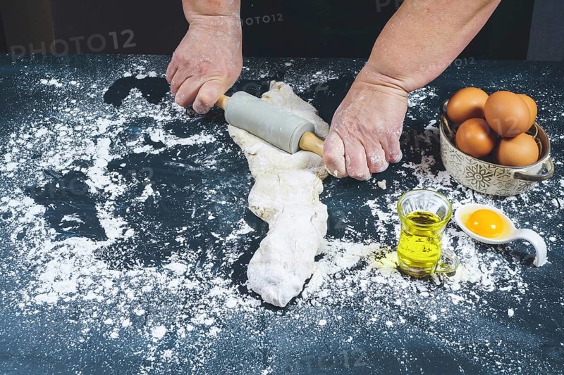 Woman kneading with a rolling pin a dough of flour to make bread on a black wooden table with olive oil