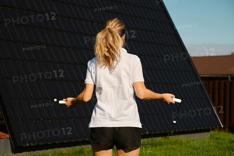 Back view of a woman jumping with a smart digital rope on the lawn in front of the house