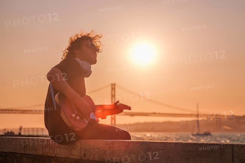 Hipster street musician in black playing electric guitar in the street on sunset on embankment with 25th of April bridge in background. Lisbon