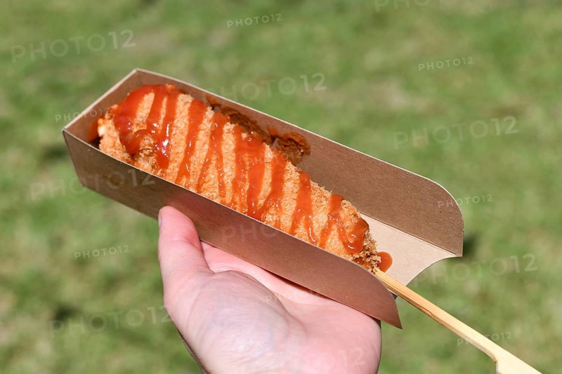 Hand holding Korean style corn dog coated in crispy panko breadcrumbs with hot sauce