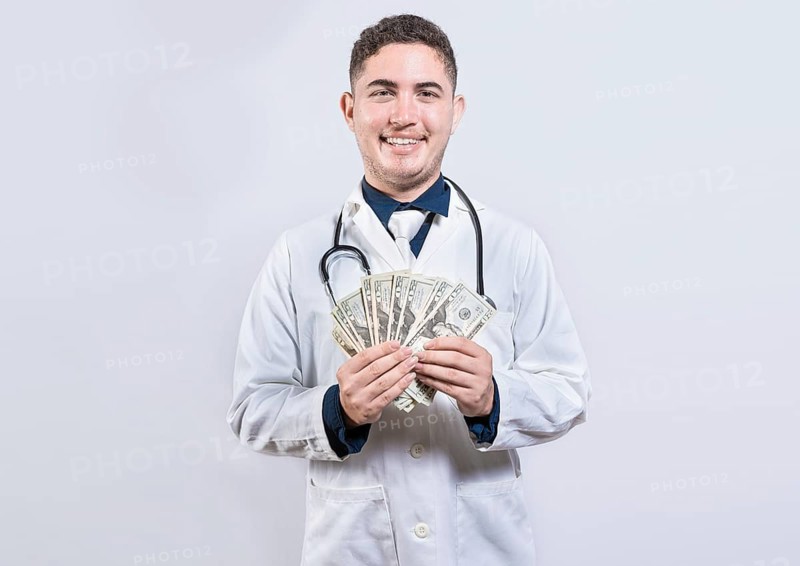 Latin young doctor holding money isolated. Cheerful doctor showing dollars isolated