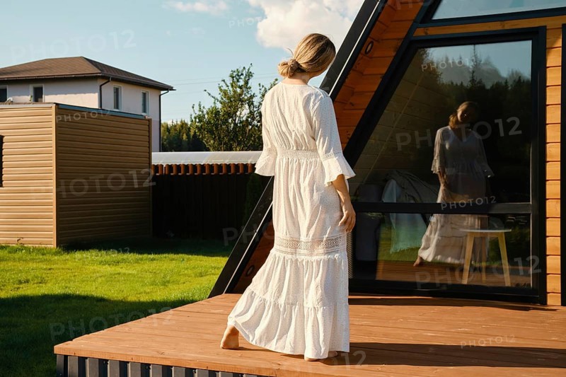 Playful woman in long white sundress looking at her reflection in the window