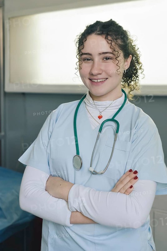 Smiling young female doctor medical nurse wearing white coat and stethoscope around neck standing in private clinic with arms crossed looking at camera. Vertical portrait