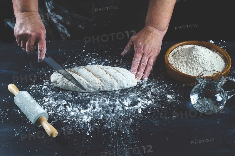 Woman cutting with a knife a flour dough to make homemade bread on a black wooden table with flour