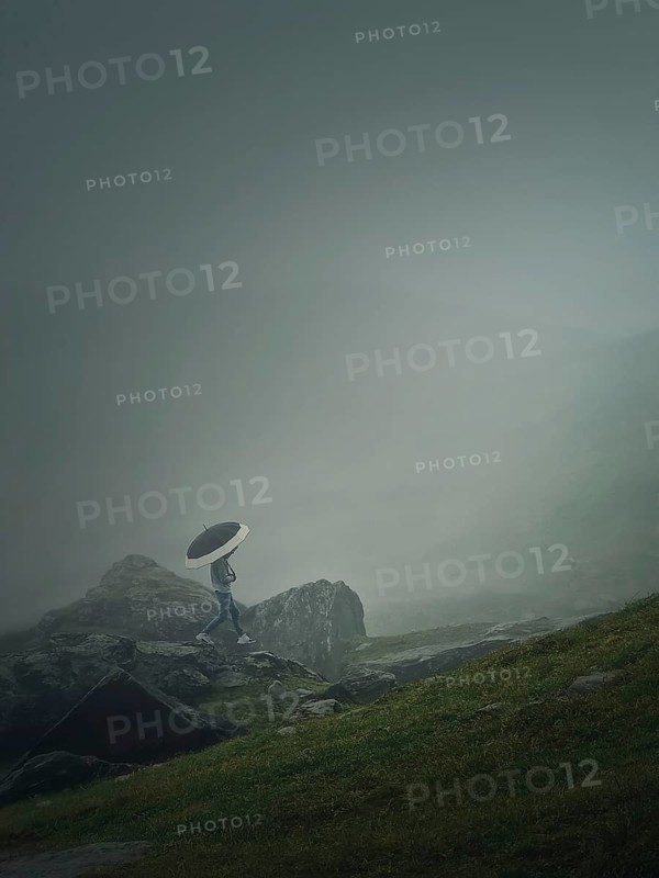 Man with umbrella climbing a mountain hill in a gloomy weather with dense mist. Moody hiking scene with a wanderer walking on the cliffs of the foggy valley