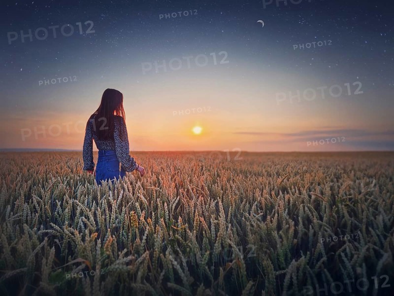 Young woman in the golden wheat field at sunset. Beautiful twilight scenery under the summer sun and crescent moon with starry sky