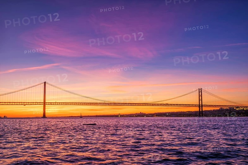 View of 25 de Abril Bridge famous tourist landmark of Lisbon connecting Lisboa and Almada over Tagus river with tourist yacht silhouette at sunset. Lisbon