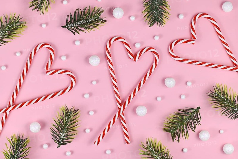 Christmas candy canes forming heart surrounded by white snowball ornaments and fir branches on pink background