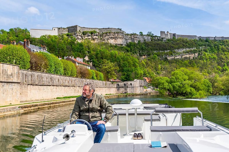 Man steering a houseboat on the river Doubs in front of the World Heritage Site of the Citadel of Besancon