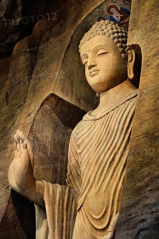 Statue of standing Buddha in monk clothes carved in limestone cave and lit by light. Right hand in adhay mudra gesture. Religion