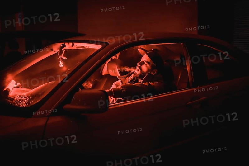 Rapper man in a cap in a car smoking at night in a garage lit with a red light in winter