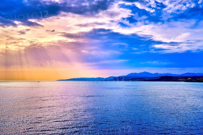 Colorful seascape with colorful clouds and bright sun rays cast over calm sea waters and hazy distant shores. Sunrise or sunset in Mediterranean sea