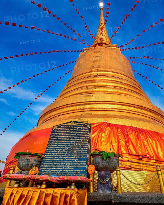 Stone board with with long message in Thai in front of big golden stupa or chedi decorated with festive red flags and textile in sunset sunlight in Wat Saket or Golden Mount temple