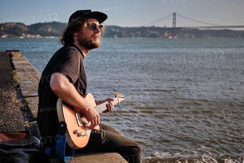 Hipster street musician in black playing electric guitar in the street sitting on pier embankment on sunset in Lisbon