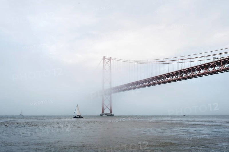 View of 25 de Abril Bridge famous tourist landmark of Lisbon connecting Lisboa and Almada in heavy fog mist wtih yacht boats passing under. Lisbon