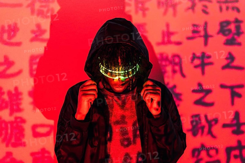 Studio portrait with red neon lights of an anonymous futuristic man wearing smart glasses hiding on a hoodie