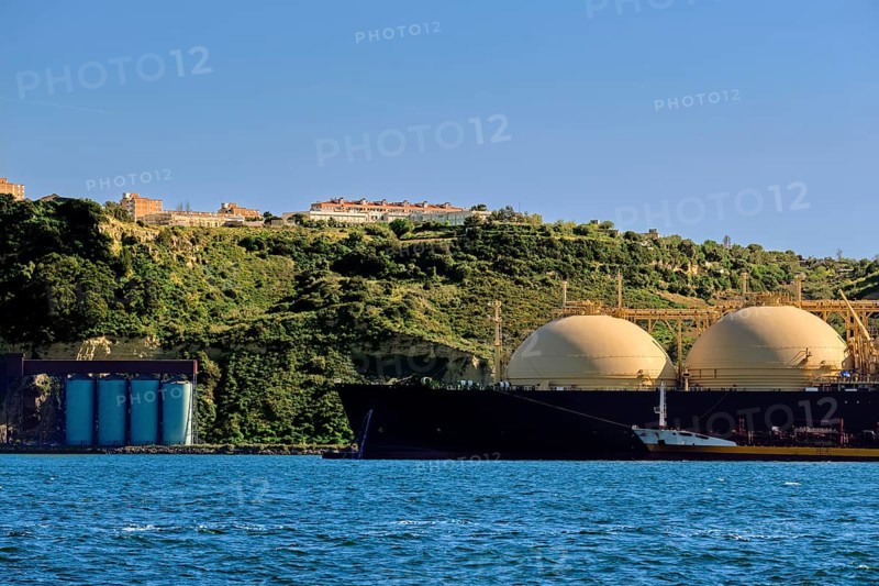 LNG or liquified natural gas tanker moored by industrial pier and green hill on sunny day. Alternative gas supply