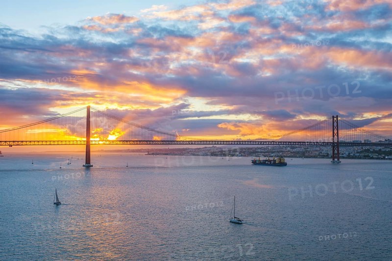 View of 25 de Abril Bridge famous tourist landmark of Lisbon over Tagus river with tourist yacht boats and cargo container ship on sunset. Lisbon