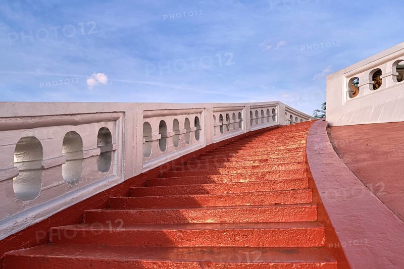 Red sunlit stairs and white banisters leading upwards at blue sky. Concept of ascension