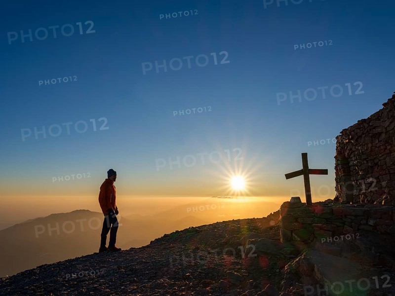 Mountaineer standing at sunset next to summit chapel Timios Stavros and summit cross of Psiloritis