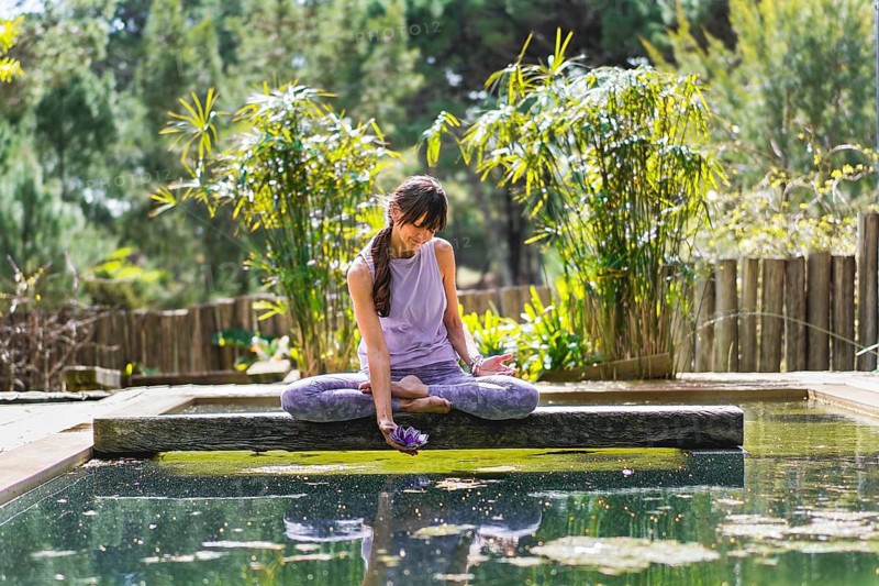 A woman holding a glass purple lotus while sitting in lotus pose on a tree trunk over the water