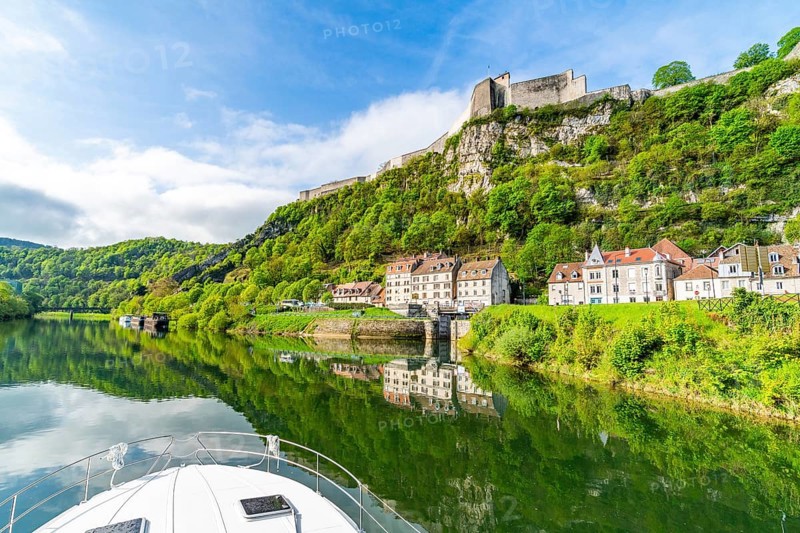 View from the bow of a houseboat on houses at the foot of the Saint Etienne mountain with the world heritage site of the citadel of Besancon on the river Doubs