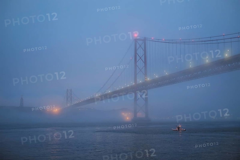 View of 25 de Abril Bridge famous tourist landmark of Lisbon connecting Lisboa and Almada in heavy fog mist wtih yacht boats passing under. Lisbon
