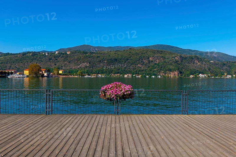 Elevated Walkway with Flowers on the Railing on Lake Lugano with Mountain in a Sunny Summer Day in Porto Ceresio
