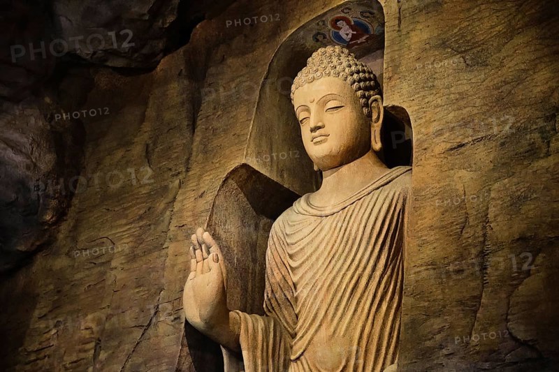 Statue of standing Buddha in monk clothes carved in limestone cave and lit by light. Right hand in adhay mudra gesture. Religion