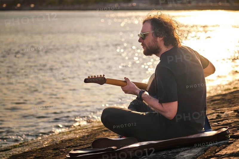Hipster street musician in black playing electric guitar in the street sitting on pier embankment on sunset